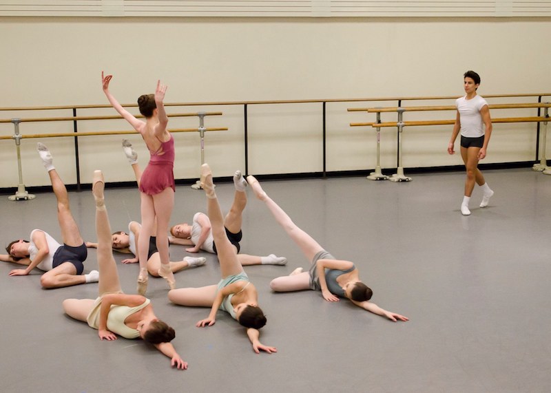 Six dancers lay on the ground with one leg in the air. A female soloist stands on pointe while a male soloist stands across from her.